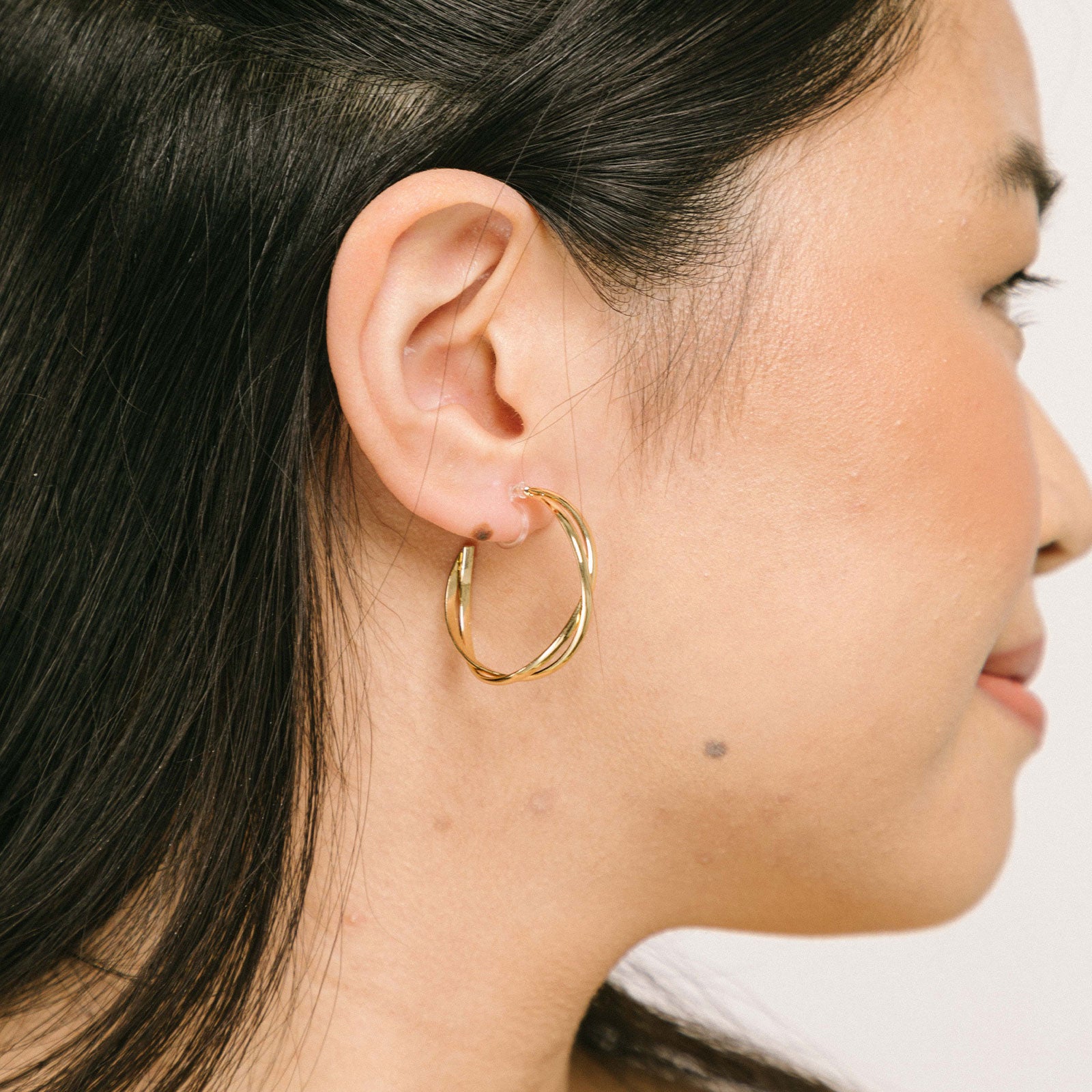 A model wearing the Gold Vienna Hoop Clip-On Earrings feature a resin clip-on closure with medium secure hold. The earrings are suitable for all types of ears, with an average comfortable wear time of 8-12 hours. No adjustments can be made. Constructed with gold tone metal alloy, these earrings are also available in silver.