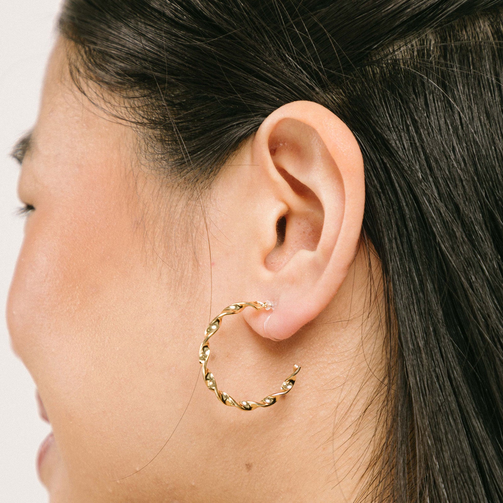 A model wearing the Gold Wave Hoop Clip-On Earrings boast a closure type of resin clip-on, ideal for the vast majority of ear types. Average wear duration ranges from 8-12 hours, offering medium secure hold with no ability to adjust. Crafted from gold tone metal alloy, this item comes as one pair and is also available in silver.