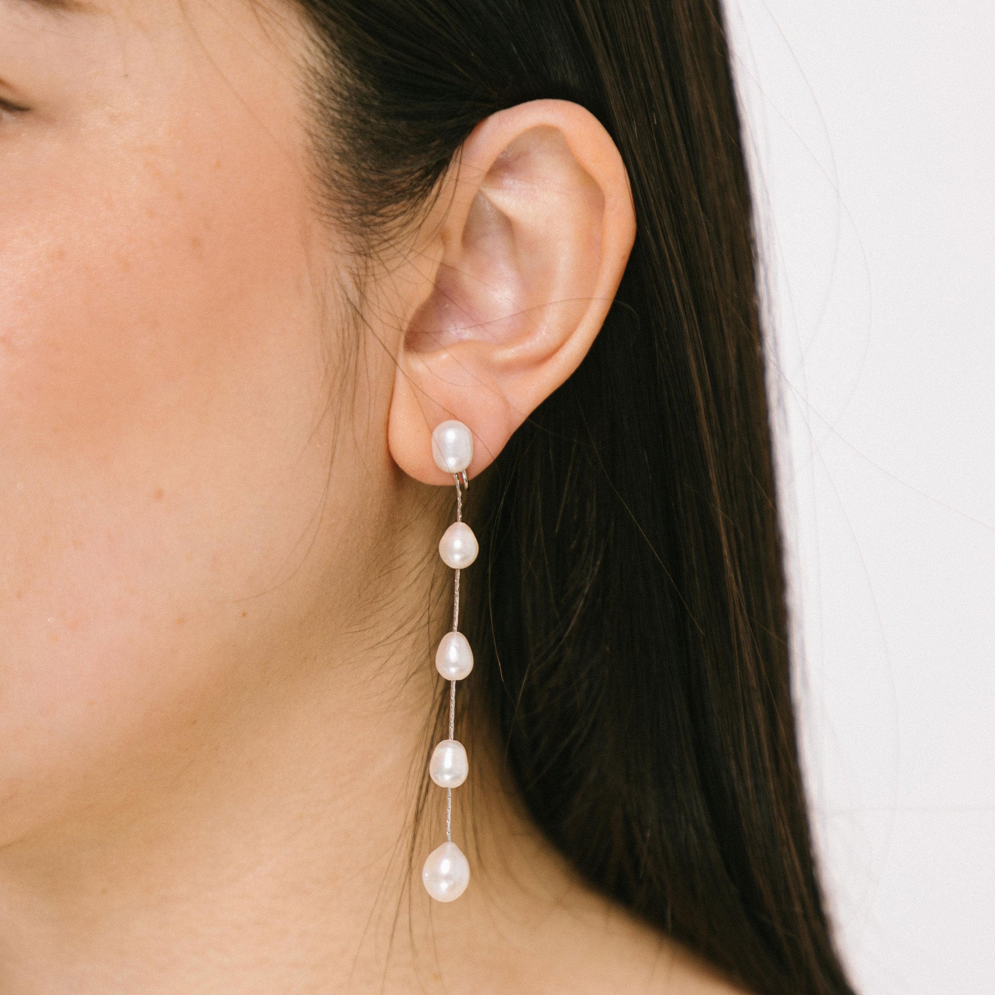A model wearing the  Lune Pearl Clip-On Earrings in Silver with ease and comfort. Their Mosquito Coil Clip-Ons feature a medium secure hold that can be adjusted to any ear size, shape, and sensitivity-level. Crafted from Freshwater Pearls, each set exhibits its own unique color and size for a one-of-a-kind look. Long-lasting design ensures it can be worn for up to 24 hours with ease.