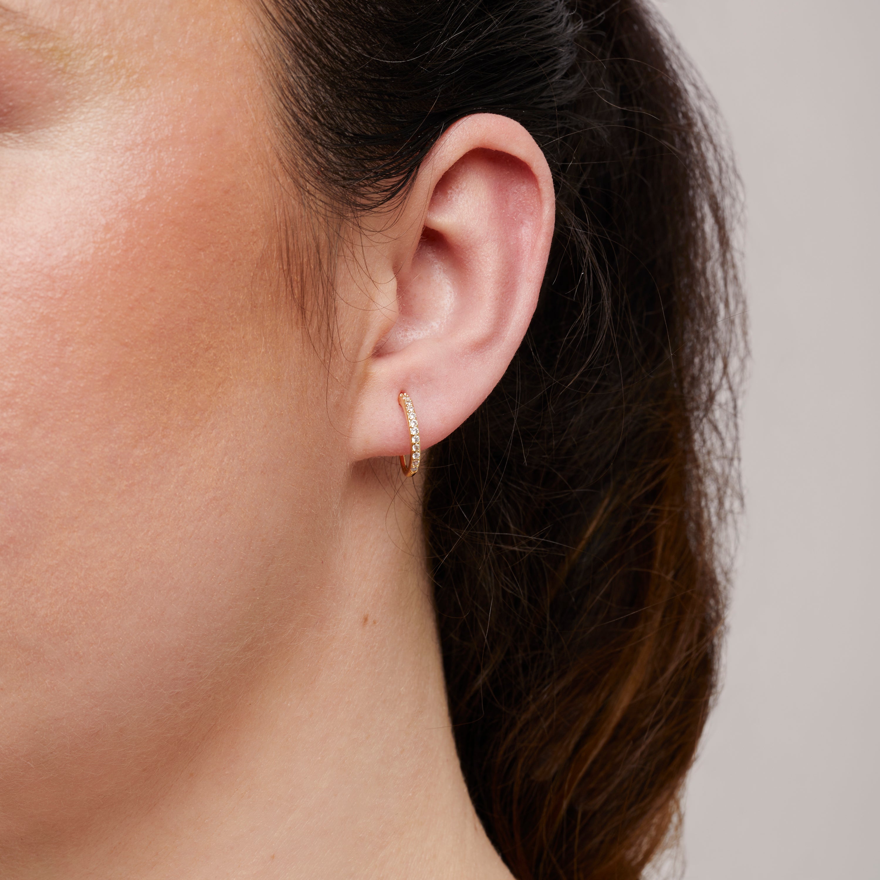 A model wearing the Medium Pavé Hoop Clip On Earrings in Gold offer a Mosquito Coil Clip-On Closure, making them ideal for Thick/Large Ears, Sensitive Ears, Small/Thin Ears, Stretched/Healing Ears, and Keloid Prone Ears. They provide an average comfortable wear duration of 24 hours with a medium secure hold, and can be gently adjusted by squeezing the padding forward once on the ear. Please note that this item is only one pair.