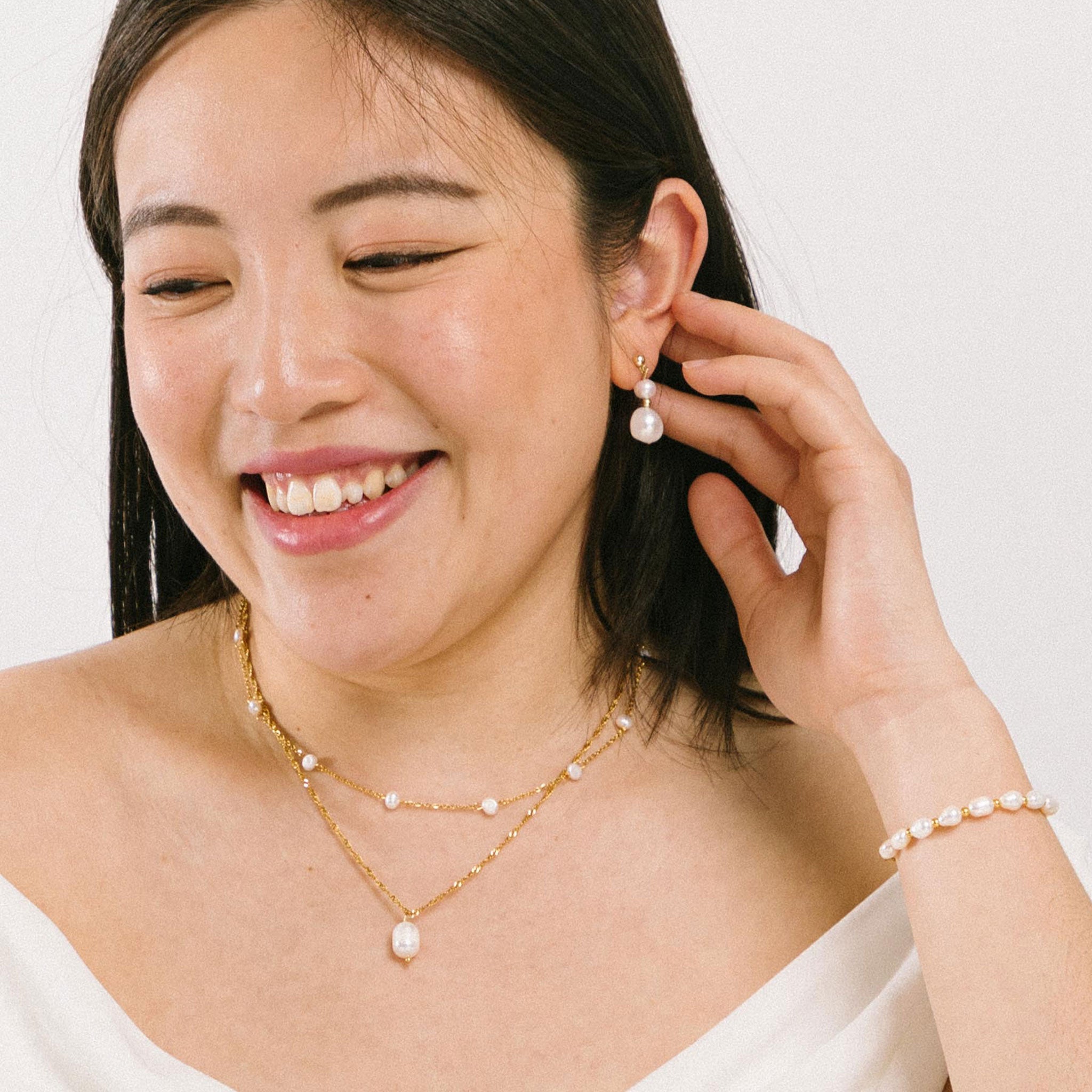 A model wearing the Elena Pearl Necklace is adjustable and crafted from Freshwater Pearls, 18K Gold Plated Stainless Steel, and is waterproof and hypoallergenic. Each pearl may vary slightly in size and colour due to its natural characteristics.