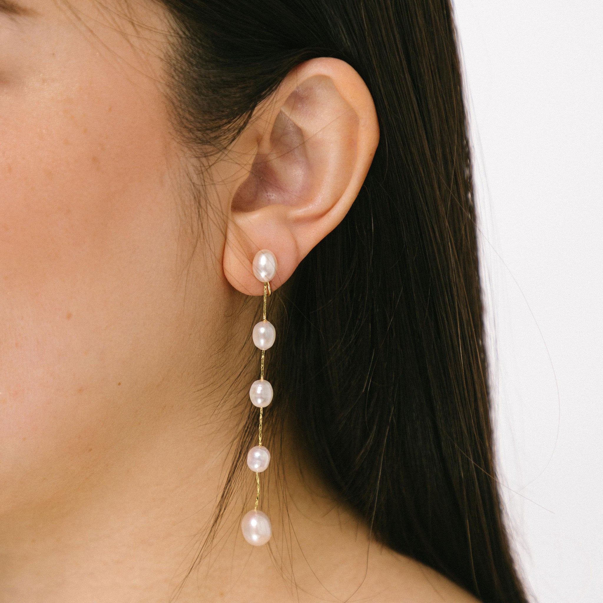 A model wearing the Lune Pearl Clip-On Earrings in Gold feature a unique, secure mosquito coil clasp. They are ideal for all ear types, from thick and large ears to sensitive and small. With medium hold strength, the earrings can be comfortably worn up to 24 hours and easily adjusted. Crafted with freshwater pearls, each pair may display natural variations in size and color. Materials include freshwater pearl and 18K gold-plated stainless steel.