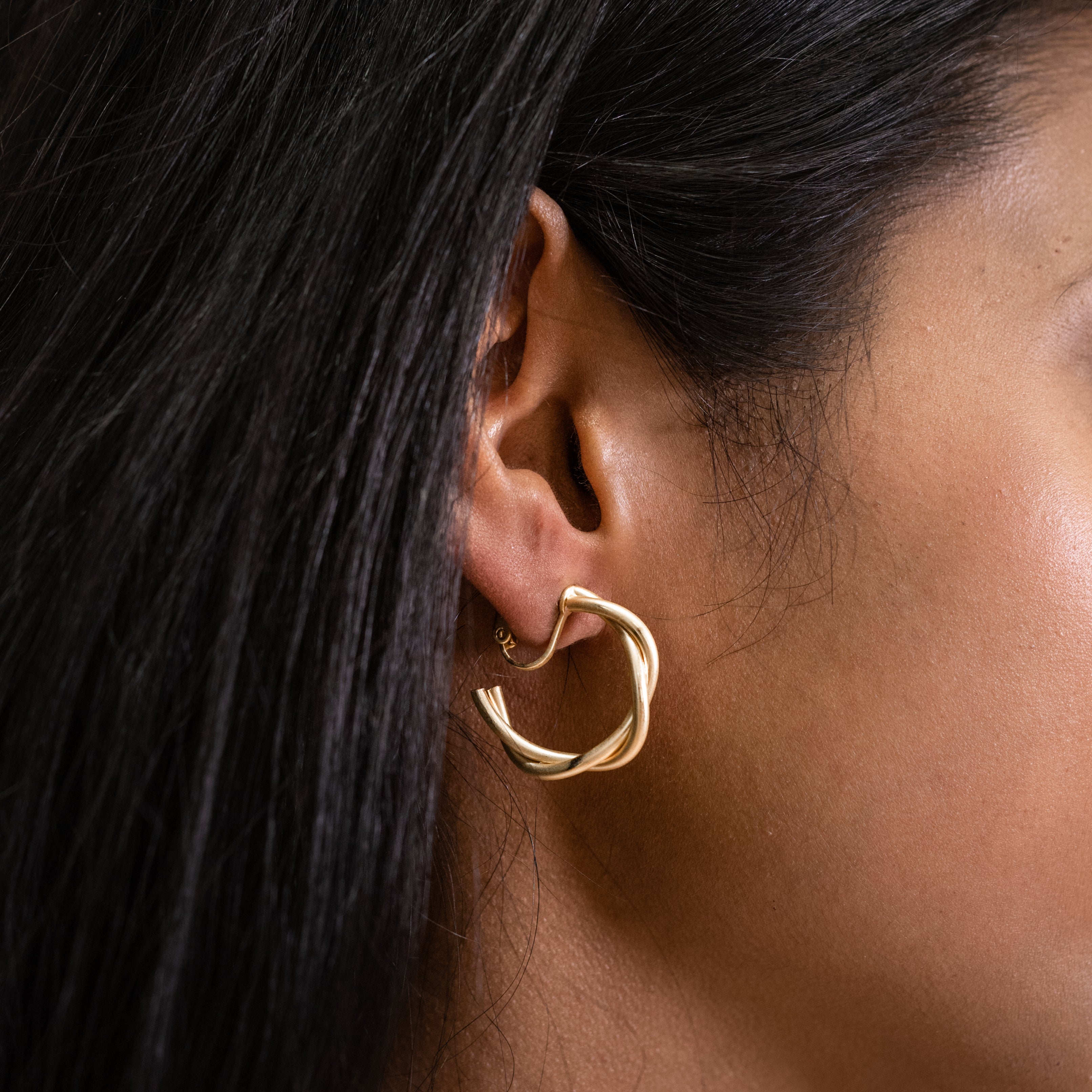 A model wearing the Twist Hoop Earrings feature a secure and comfortable screwback clip-on closure ideal for all ear types. Wear these earrings for up to 8-12 hours per day with a secure hold and the ability to be manually adjusted to the individual's size. Crafted from gold tone plated copper alloy, these earrings come is a single pair.